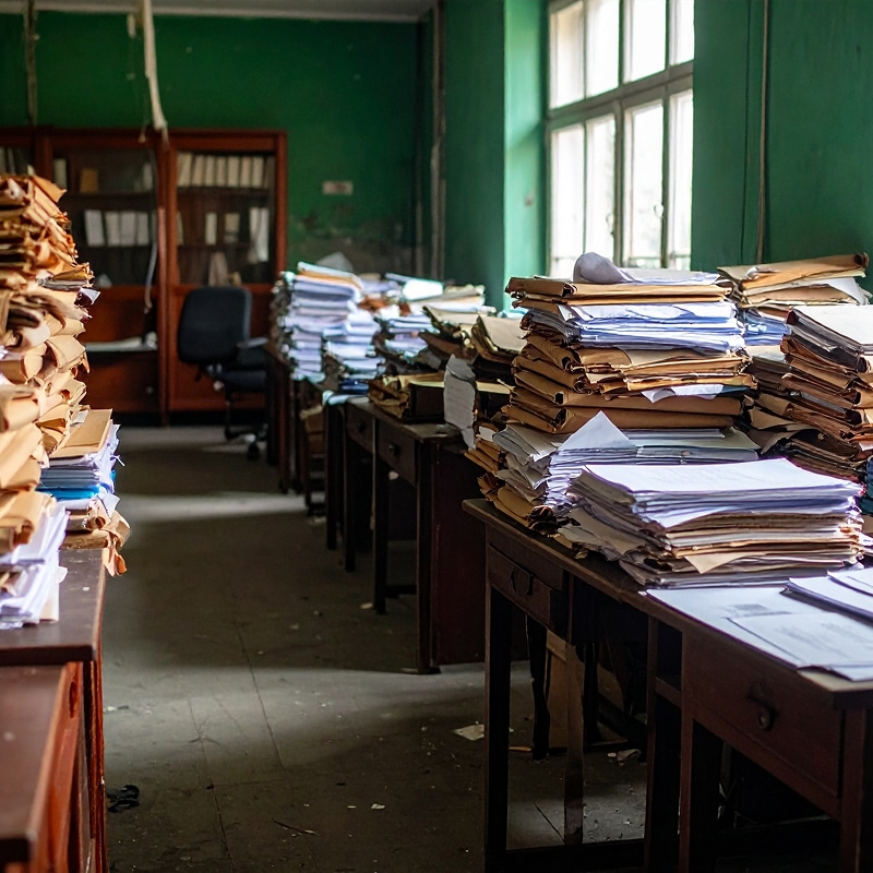 Bureau ancien encombré de piles massives de dossiers jaunis et de papiers empilés sur des bureaux en bois sombres, murs vert émeraude.