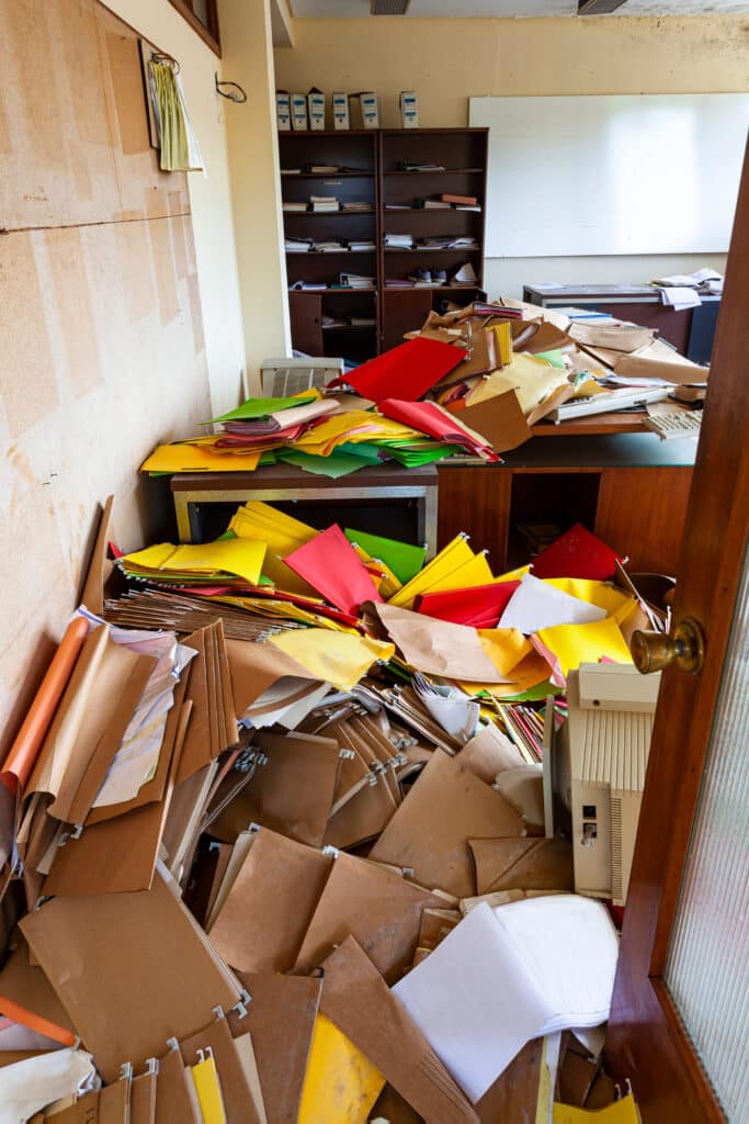 Bureau envahi par des piles de dossiers suspendus marron, jaunes, rouges et verts, vu par une porte ouverte.
