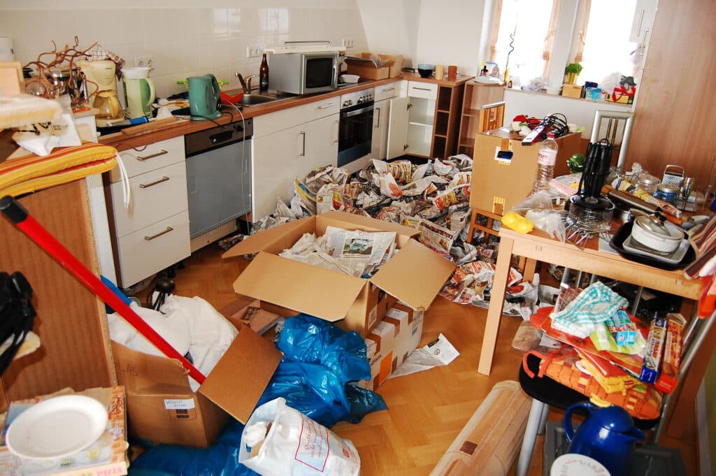 Cuisine très désordonnée avec cartons de déménagement, sacs bleus, journaux étalés et vaisselle éparpillée sur les plans de travail et le sol en parquet.