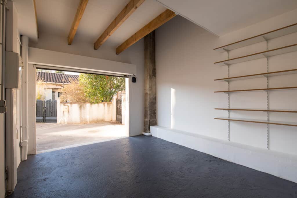 Garage vide avec poutres bois et étagères murales Intérieur de garage vide avec sol gris foncé, poutres en bois apparentes et étagères murales blanches, vue sur l'extérieur ensoleillé par la porte ouverte.