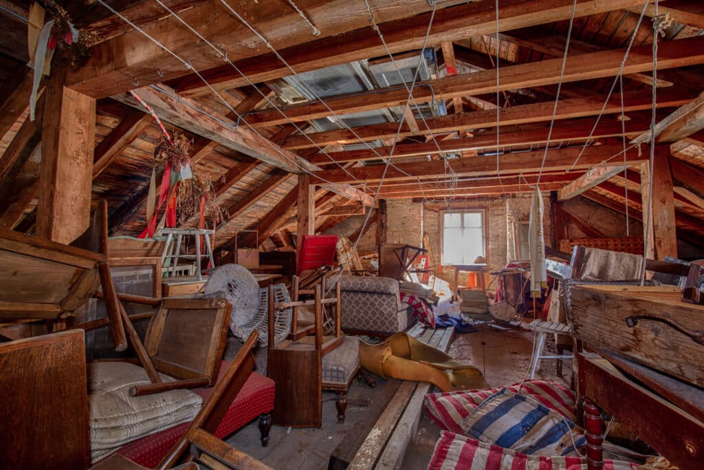 Grenier poussiéreux et encombré, éclairé par une fenêtre, rempli de meubles anciens renversés, de bois apparent et de cordes.